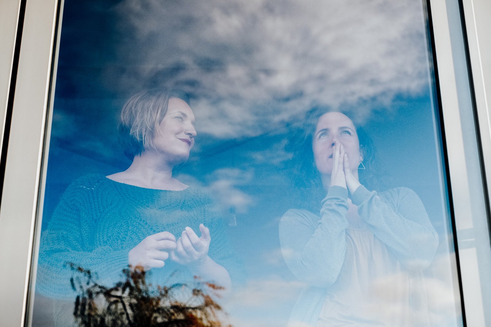 Twee vrouwen achter een raam met wolken erin weerspiegeld, in reflectie en verbinding